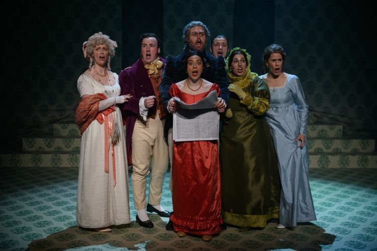 Cast of Mansfield Park looking shocked with the central cast member reading a newspaper. All are dressed in flamboyant colourful Victoria style costumes.